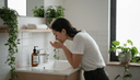 Jeune femme de profil se rinçant le visage au-dessus d'un lavabo, flacon nettoyant et serviette pliée