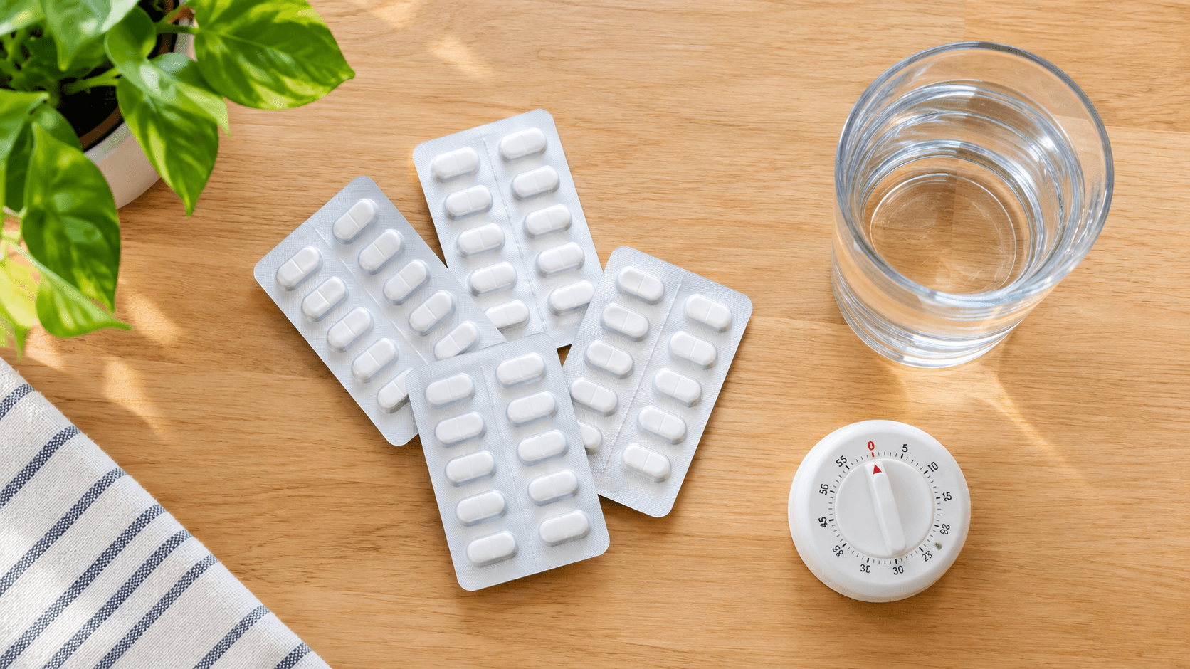 Vue de dessus de plaquettes de comprimés blancs, un verre d'eau et un minuteur posés sur une table en bois clair près d'une plante