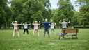 Groupe de sexagénaires de dos pratiquant le tai-chi sur une pelouse de parc, tapis sur un banc