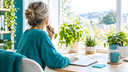 Femme d'une cinquantaine d'années de dos à un bureau lumineux, notant une pensée, entourée de plantes vertes