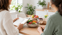 Deux adultes de profil discutent autour d’une table en bois avec un carnet, des légumes, des lentilles et une carafe d’eau