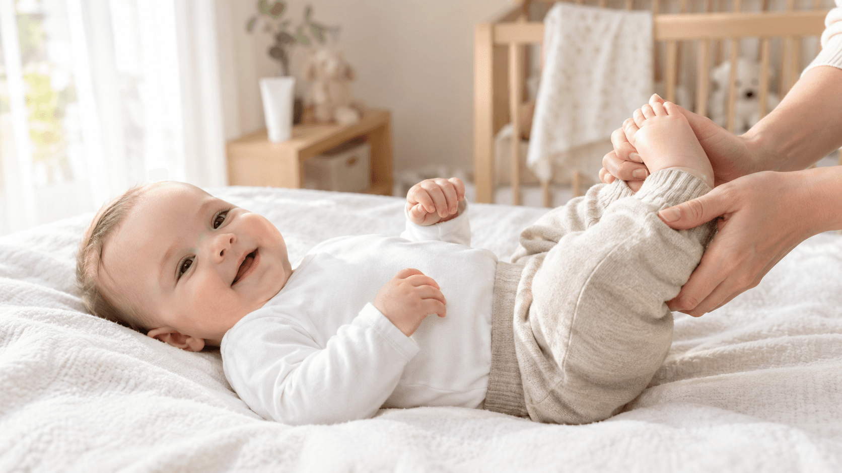 Bébé souriant vêtu de coton blanc sur une couverture, les mains d’un parent tenant ses pieds dans une chambre lumineuse