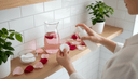 Vue plongeante de mains vaporisant un hydrolat sur un coton, pétales de rose et carafe