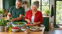 Couple de sexagénaires souriants préparant un repas méditerranéen de légumes, lentilles et sardines en cuisine lumineuse