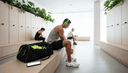 Sportif vu de dos laçant ses chaussures sur un banc, un casque audio autour du cou et un sac ouvert