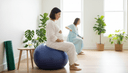 Une femme enceinte assise de profil sur un ballon, mains sur son ventre, tapis roulé et verre d'eau