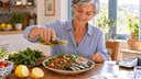 Femme quinquagénaire versant de l'huile d'olive sur un plat de sardines, lentilles et légumes, tensiomètre sur la table