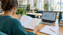 Femme vue de dos lisant un épais rapport scientifique annoté dans une bibliothèque lumineuse, carnet et ordinateur à côté